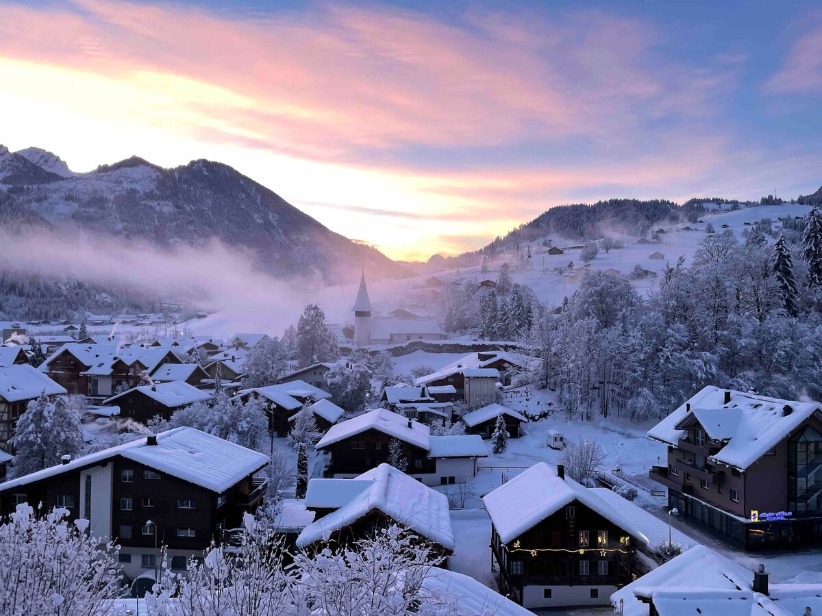 A snowy landscape captures a charming village beneath a pastel sky at sunrise. Snow-covered rooftops are visible, along with distant mountains and a church steeple peeking above the trees. The serene scene reflects the tranquility of a winter day in the region.