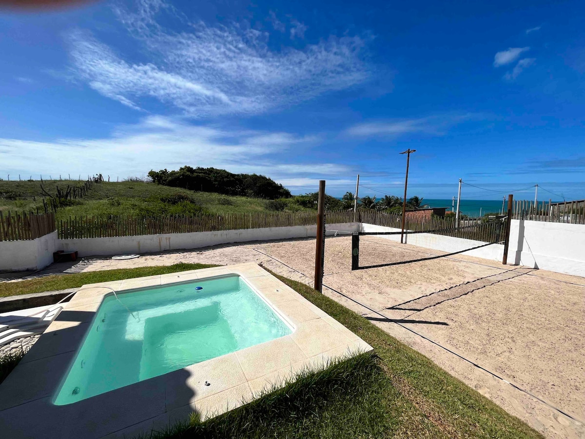 A spacious outdoor area features a pool spa with a clear blue water surface, complemented by a sand court for beach tennis and volleyball. Lush green hills are visible in the background, with a bright blue sky overhead, creating a serene environment.