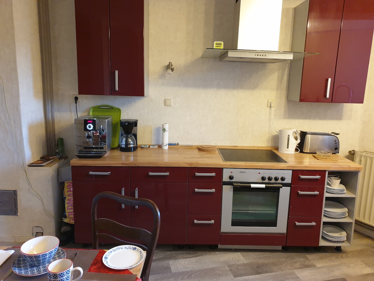 A modern kitchen features red cabinetry and a wooden countertop. Appliances include a coffee maker, kettle, microwave, and oven. The setup allows for meal preparation, with dishes and mugs visible on the table nearby. Flooring is light-colored and complements the overall aesthetic.
