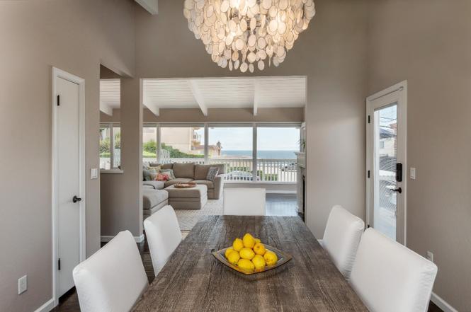 A dining area features a large wooden table with a bowl of fresh lemons at its center. Light-colored chairs surround the table, and large windows allow natural light to fill the space while offering views of the ocean beyond.
