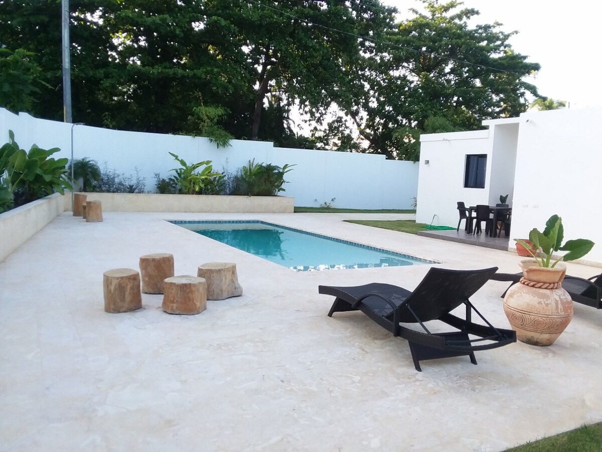 A private pool is surrounded by a spacious patio, featuring wooden stools and reclining lounge chairs. Lush green plants enhance the outdoor area, while a white wall provides privacy. A dining table can be seen in the background, creating a serene setting for relaxation.