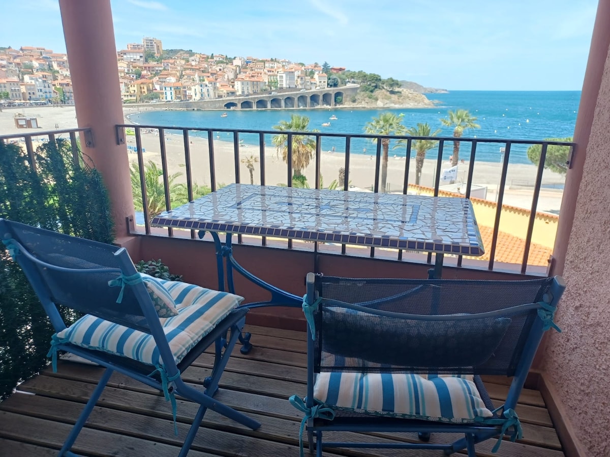 A balcony features two striped chairs and a table, overlooking a view of the beach and coastline. In the distance, colorful buildings line the shore, while palm trees are visible along the sandy beach, contributing to a relaxed seaside ambiance.