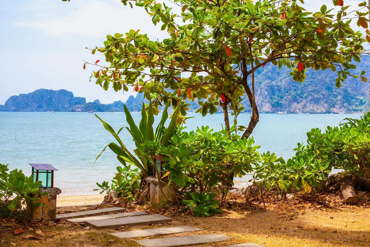 A pathway of stone slabs leads toward the calm shoreline, framed by lush greenery and a scattering of rocks. A small table is positioned nearby under a leafy tree, offering tranquil views of the water and distant mountains.