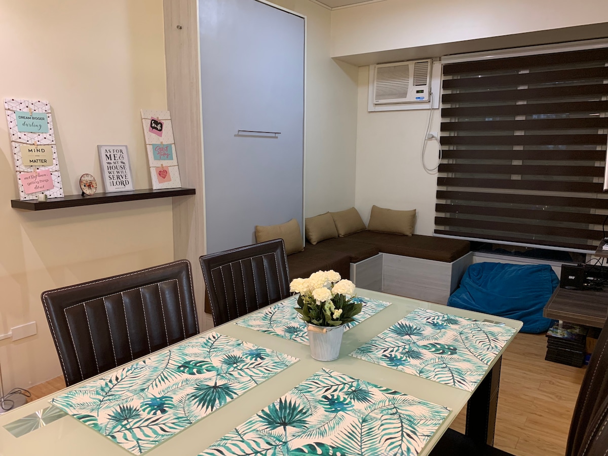 A dining area features a glass-topped table adorned with tropical-themed placemats, surrounded by dark wooden chairs. A small shelf displays decorative items and framed artwork. A cozy seating nook is positioned by the window, with cushions against the wall and a blue bean bag on the floor.