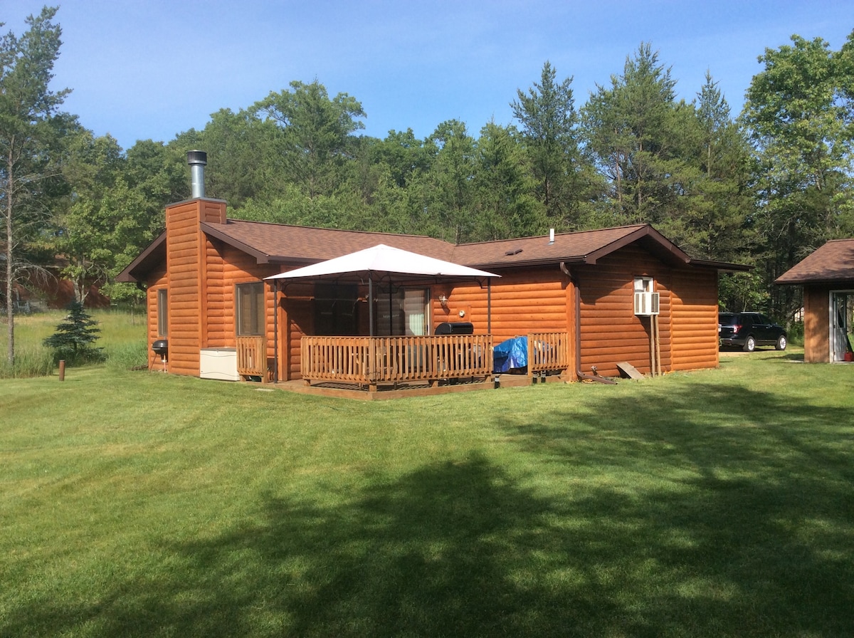 A wooden cabin with a sloped roof is set amidst a grassy area. A shaded deck with a railing extends from one side, while trees surround the property, offering a natural backdrop. A chimney indicates heating capabilities, and additional structures are visible in the distance.