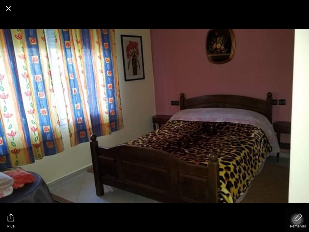 A simple bedroom is presented with a wooden bed featuring a patterned blanket. Colorful curtains frame a window, allowing natural light to fill the space. Walls are painted in soft tones, complemented by framed artwork that adds character to the room.