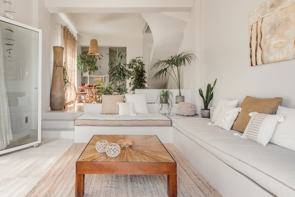 A light-filled lounge area features a spacious built-in seating arrangement adorned with neutral-toned cushions. A wooden coffee table with decorative items sits in the center, while potted plants provide a touch of greenery. The overall design reflects a modern yet relaxed ambiance.