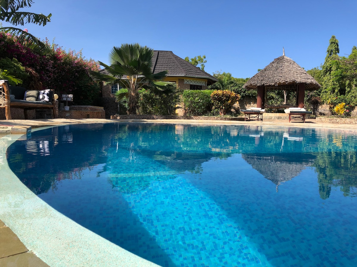 A clear blue pool reflects the surroundings, with a thatched gazebo and two Lamu beds in the background. Lush greenery and vibrant flowers frame the space, creating a serene and inviting outdoor atmosphere perfect for relaxation.