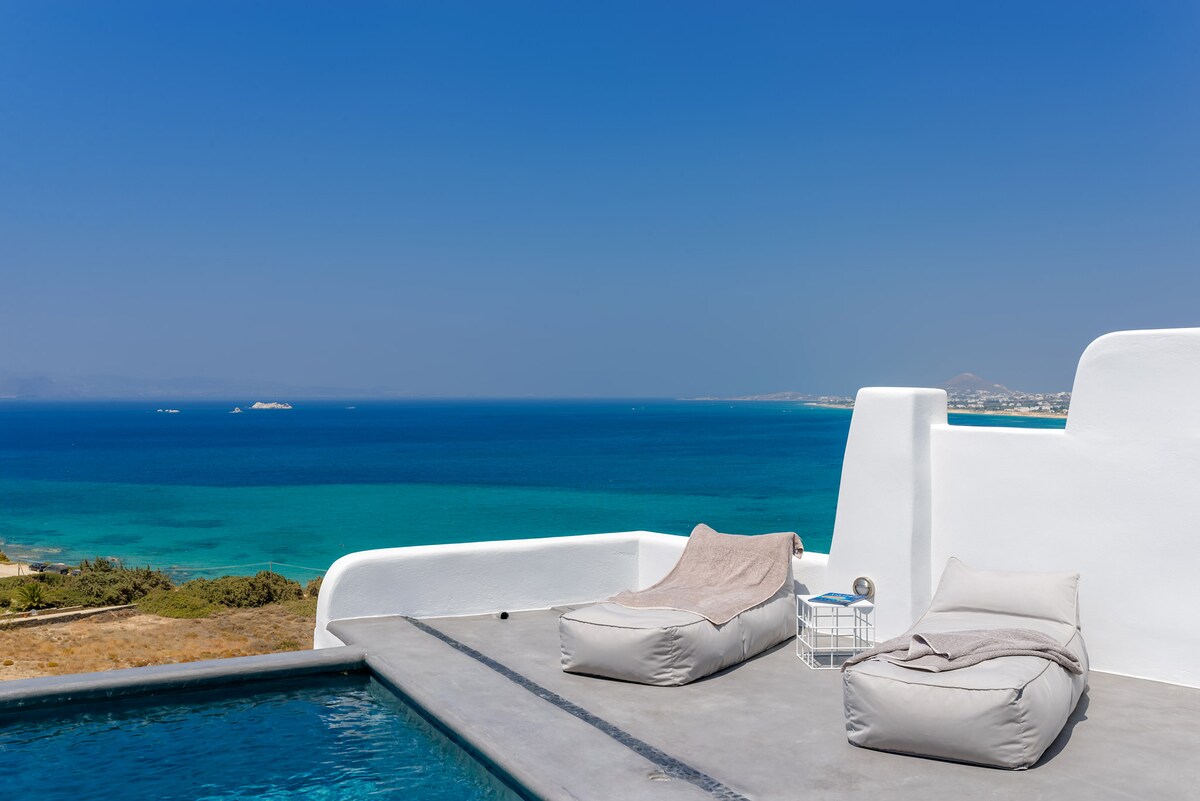 A rooftop terrace is shown overlooking the crystal-clear sea. Two plush loungers are positioned near the edge, providing comfort while enjoying the view. A small round table accompanies them, completing the relaxing outdoor space under a clear blue sky.
