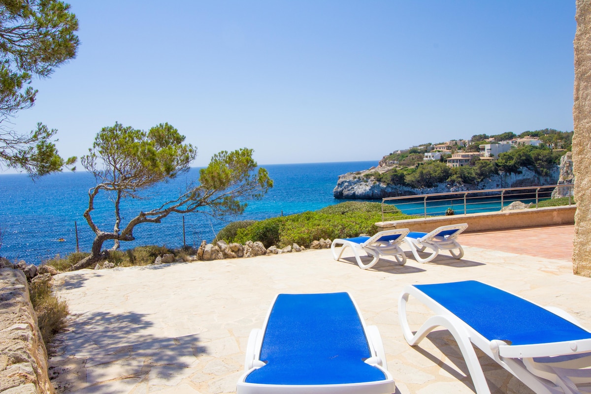A serene terrace area features multiple sun loungers positioned on a stone floor, overlooking the sparkling Mediterranean Sea. Lush greenery is visible in the foreground, while a cliffside landscape can be seen in the distance against a clear blue sky.