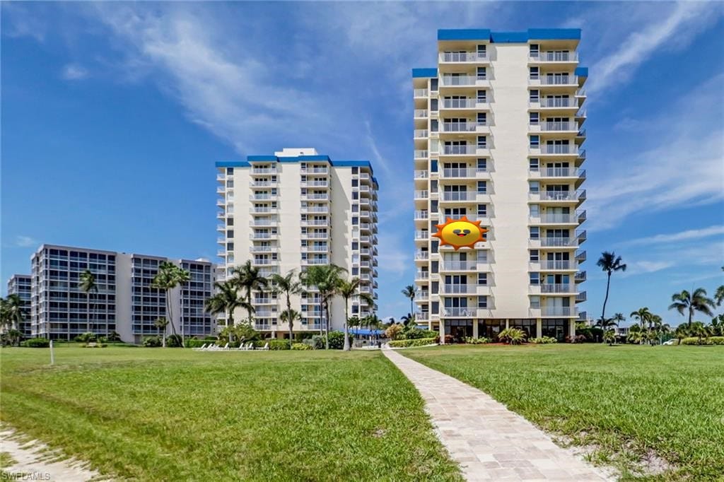 Two tall condominium buildings are visible, set against a bright blue sky. A neatly paved pathway leads through well-maintained grassy areas, connecting the buildings. Lush greenery surrounds the path, adding to the inviting atmosphere of the beachfront property.