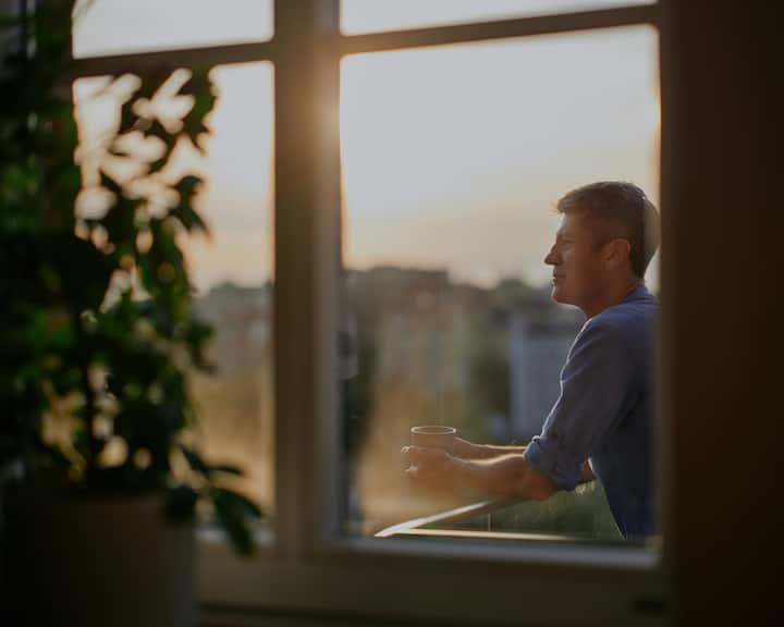 Un bărbat în cămașă albastră ține în mână o cană de cafea, în timp ce stă în picioare pe balcon și privește în zare.