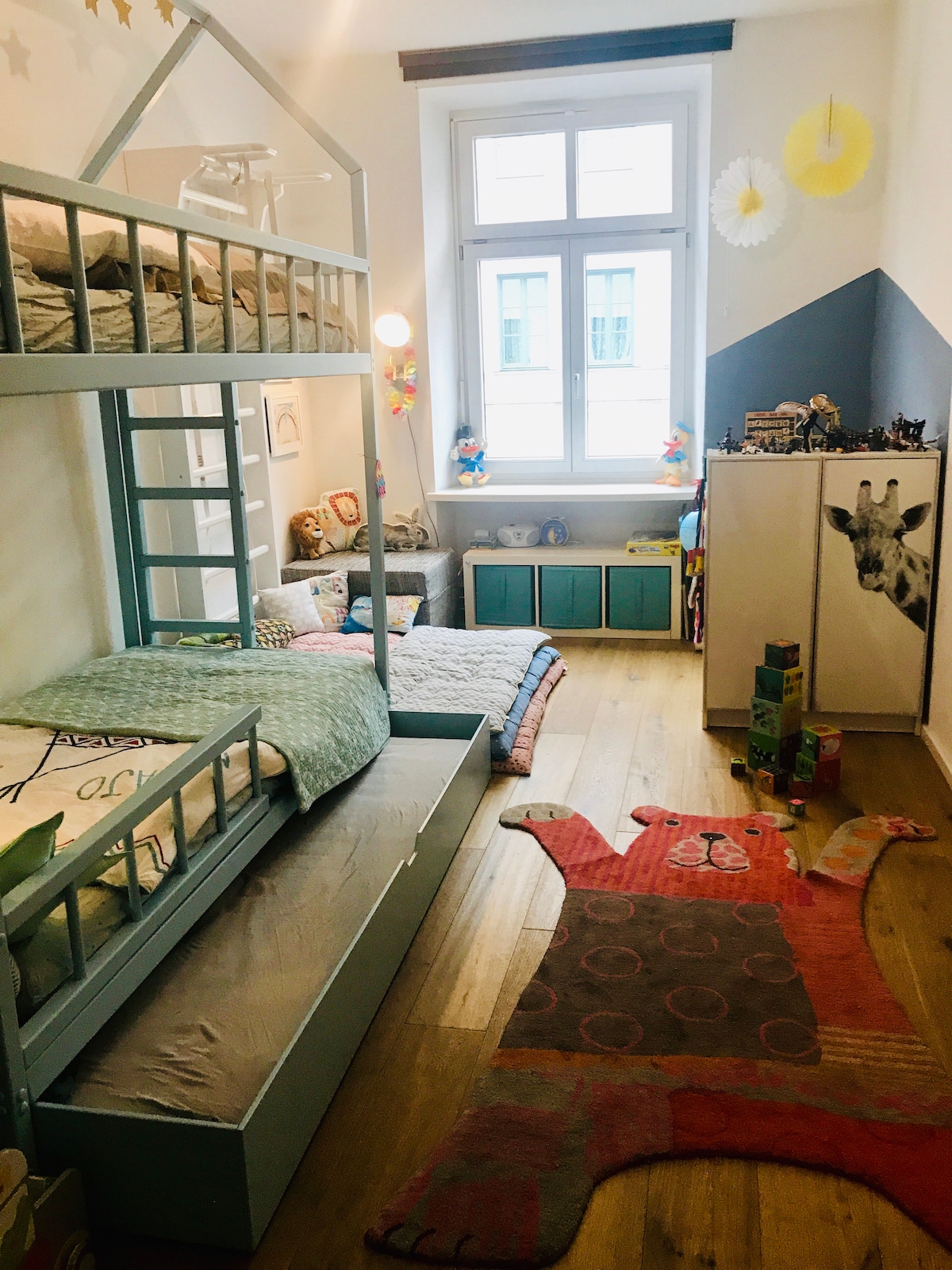 A children's bedroom features a light blue bunk bed with a loft bed above and a lower sleeping area. A colorful bear rug adds warmth to the wooden floor, and a large window provides natural light. Shelves are filled with toys and decorative items.