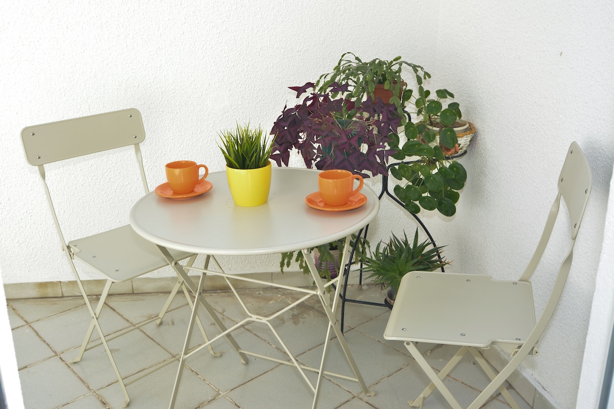A small terrace is featured with a round table and two folding chairs. It is adorned with a vibrant yellow planter and several small potted plants. Two orange cups are placed on the table, adding a pop of color to the tranquil setting.