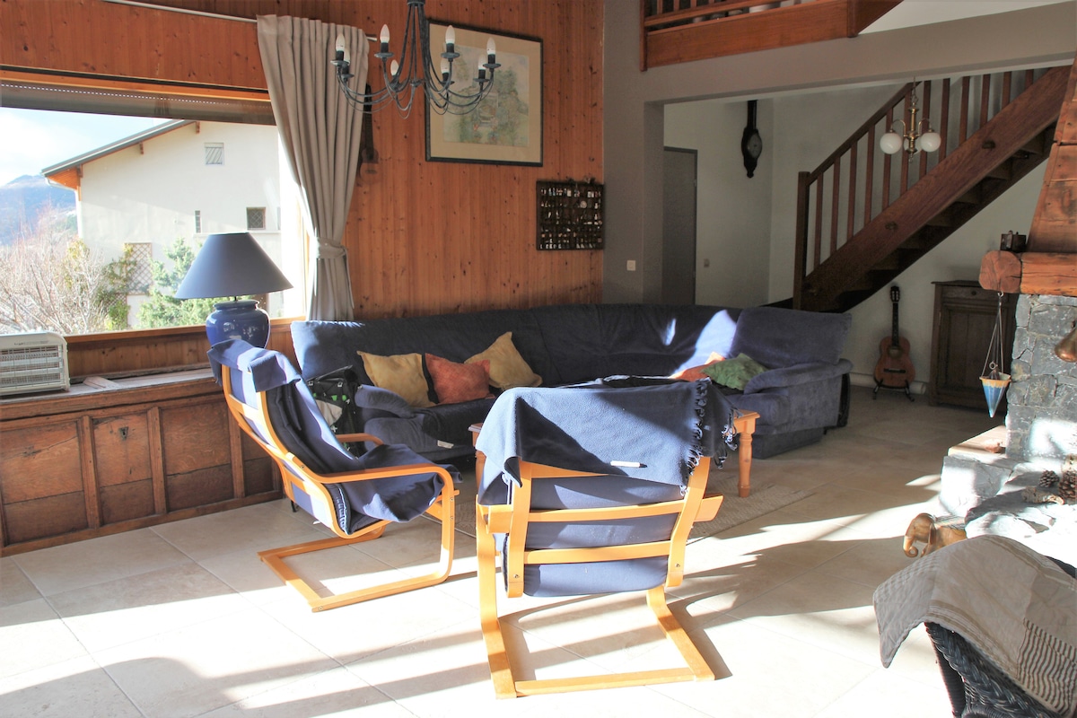 A spacious living area is highlighted by a large sectional sofa adorned with colorful cushions. A wooden staircase leads to the upper level, while a soft light streams through a window. A cozy armchair and a decorative lamp complete the inviting space.