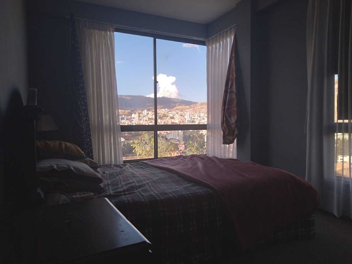 A spacious bedroom is equipped with a comfortable bed covered in a warm blanket. Large windows offer views of La Paz and are framed by light, flowing curtains. A nightstand is visible, contributing to the room's functional design.