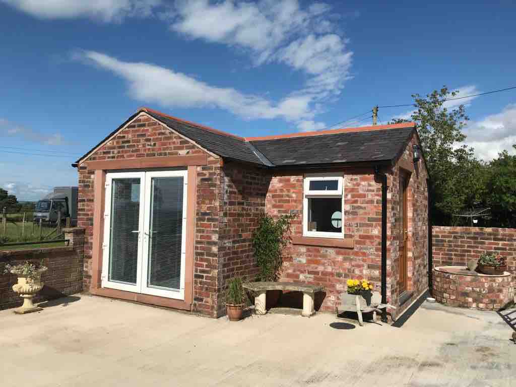 A charming guest suite with a brick exterior is set against a clear sky. Large double doors provide access to the interior, framed by a small potted plant. A stone bench and flower pots are positioned on the patio area, enhancing the outdoor space.