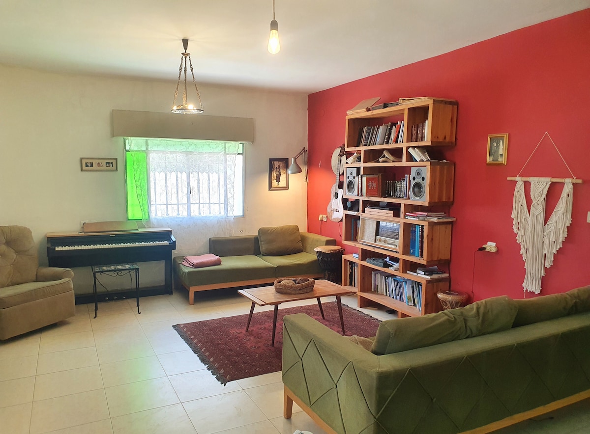 A spacious living area features a mix of comfortable seating options, including a pair of green sofas and a beige armchair. A wooden shelving unit showcases various books and a stereo. A piano is positioned near a window, with soft green curtains adding a touch of color.