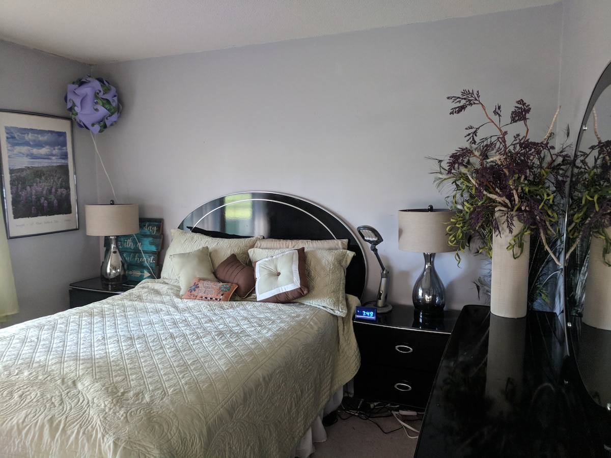 A cozy bedroom features a spacious bed with layered bedding, accompanied by decorative pillows. On either side of the bed, stylish lamps provide soft lighting. A dresser is visible, adorned with a decorative vase containing greenery. Natural light fills the room through a nearby window.