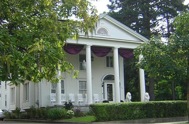The historic mansion showcases grand white pillars supporting an elegant facade. Dark fabric drapes gracefully across the entrance, framing the front door. A manicured hedge borders the property, with rocking chairs positioned on the porch, offering a welcoming space for guests to relax.