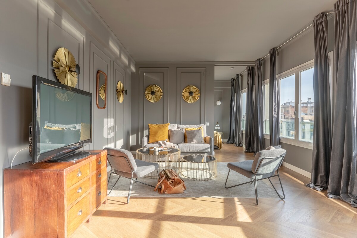 A modern living area is highlighted by large windows that allow natural light to fill the room. A cozy gray sofa and decorative cushions are complemented by stylish chairs. A sleek coffee table occupies the center, while wall-mounted decorations enhance the elegant design.