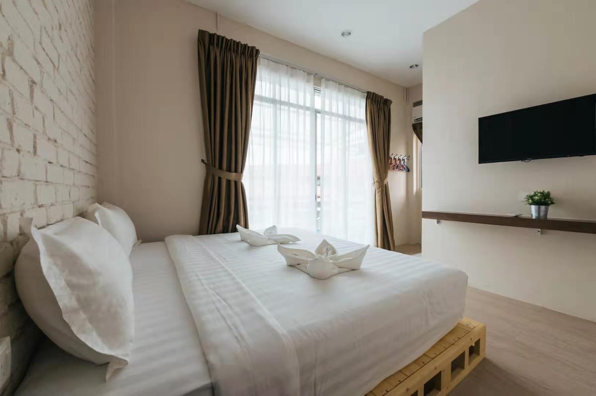 A spacious sleeping area is seen, featuring a large bed with crisp white linens and decorative towel arrangements. Natural light filters through the large window, which is framed by dark curtains. A wall-mounted television and a small shelf are positioned opposite the bed.