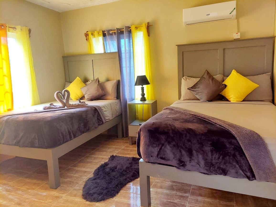 A spacious guest room is presented with two large beds, adorned with decorative yellow and gray pillows. Soft lighting from a nearby lamp creates a cozy ambiance, while light curtains allow natural light to filter in. A plush rug is positioned on the tiled floor.