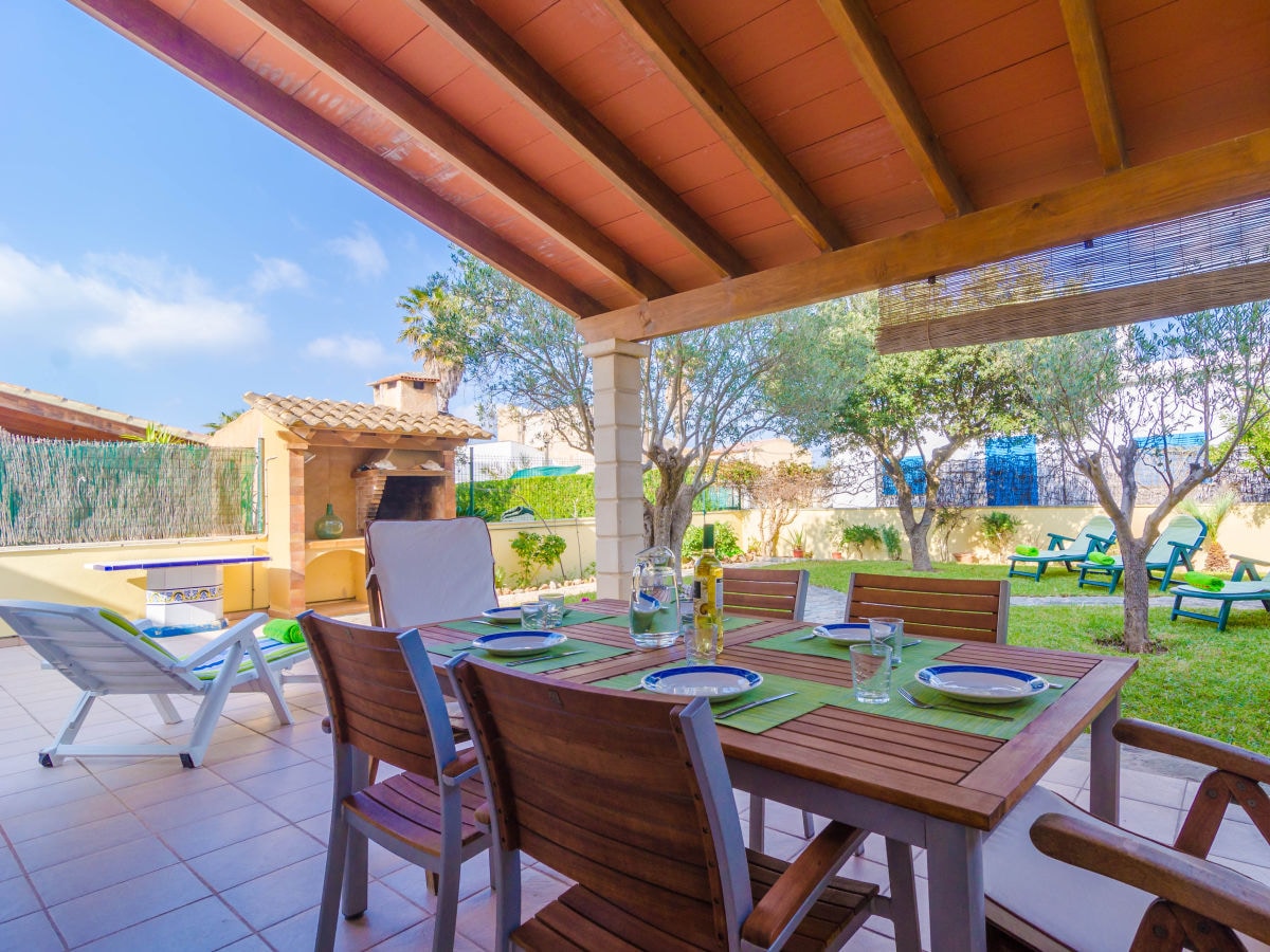 An outdoor dining area is seen under a wooden awning, featuring a table set with green placemats and tableware. A barbecue grill is positioned nearby, surrounded by well-maintained grass and trees, offering a serene setting for gatherings.