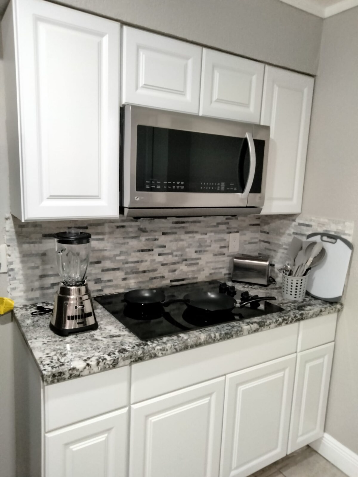 A modern kitchen area features sleek white cabinetry and a light-colored granite countertop. Appliances include a stainless steel microwave and a blender, alongside a stovetop. Utensils are neatly arranged in a holder, with a cutting board adding to the functional design.
