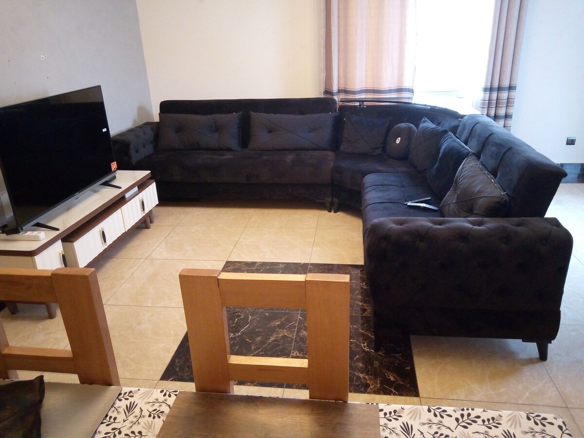 A spacious living area features a dark sectional sofa arranged around a low television stand. A flat-screen TV is positioned on the stand, while a dining table with wooden chairs is visible in the foreground. Natural light filters in through sheer curtains.