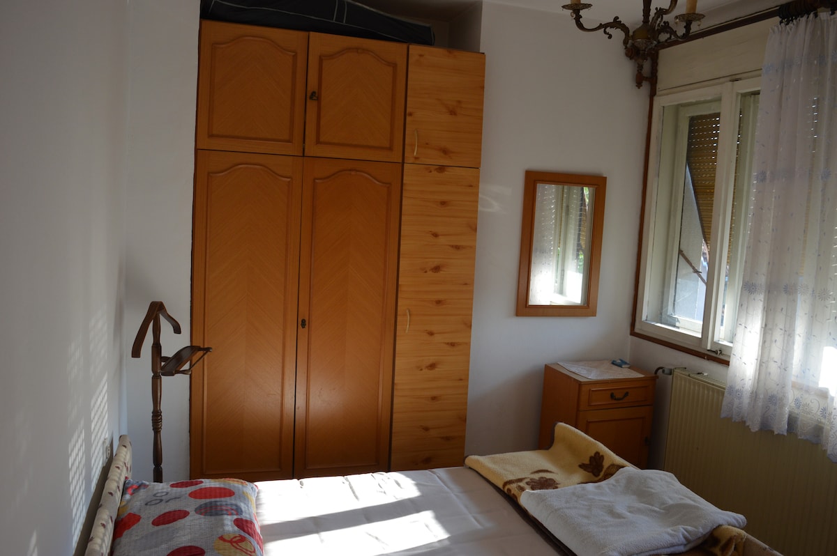 A cozy sleeping area features a spacious bed complemented by a light-colored blanket. Natural light enters through a partially open window, illuminating a wooden wardrobe and a small dresser with a mirror. A bedside lamp stands beside the bed, enhancing the room's functionality.