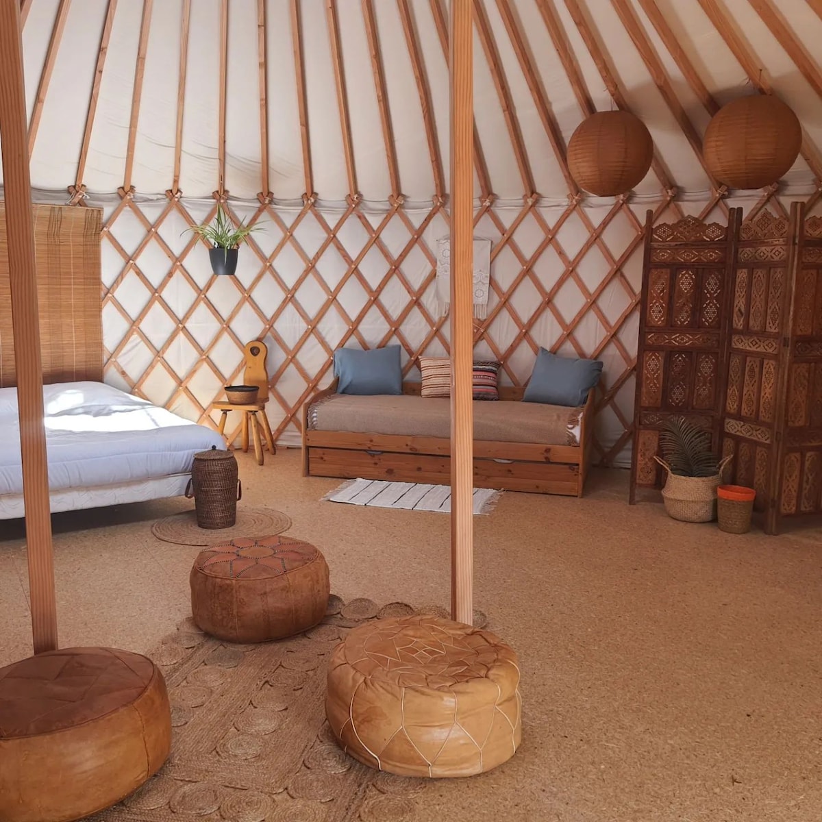 An interior of a cozy yurt is shown, featuring a spacious bed with a soft bedding arrangement. Wooden beams create a warm framework, while striped floor cushions and a decorative screen enhance the inviting atmosphere. A small table and comfortable chairs offer additional seating options.