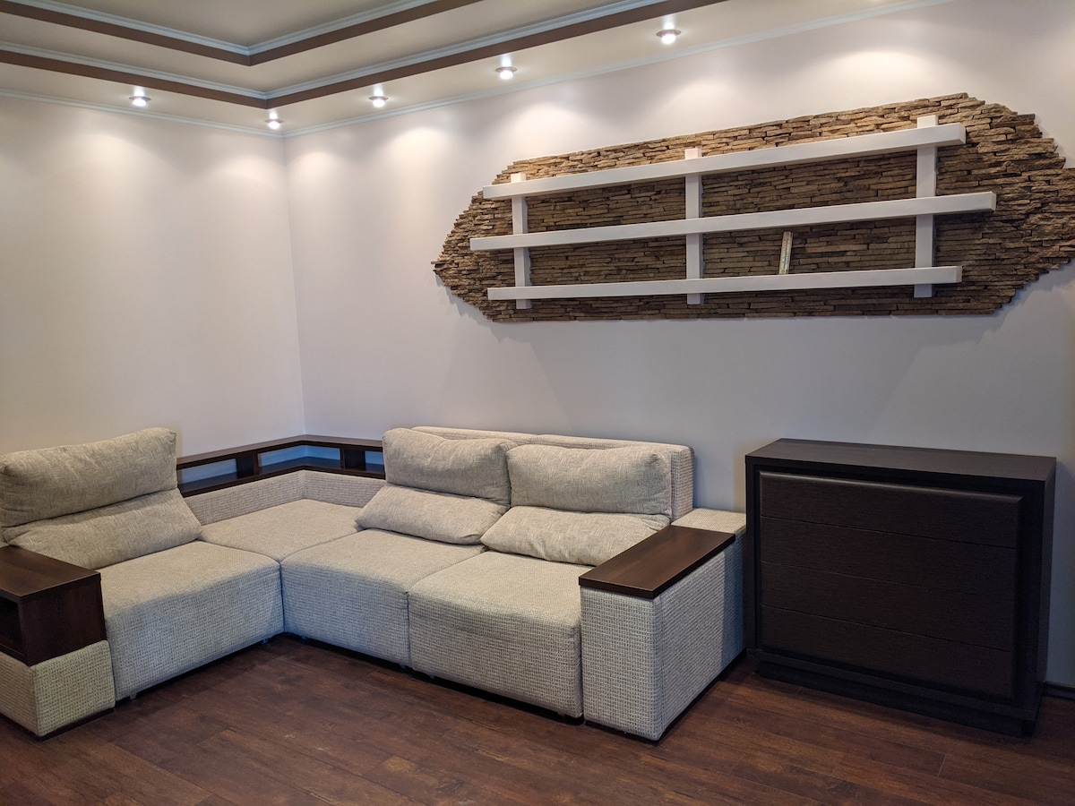 A contemporary living area is presented, featuring a sectional sofa with a light fabric finish. A sleek wooden cabinet stands against the wall, complemented by a decorative shelf made of textured stone. Soft lighting enhances the clean lines of the space.