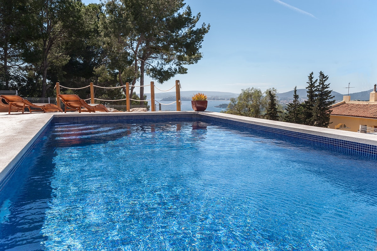 A tranquil salt-water pool glistens under the sun, surrounded by sun loungers. The scenic backdrop includes distant sea and mountain views, framed by lush greenery and trees, enhancing the outdoor relaxation space.