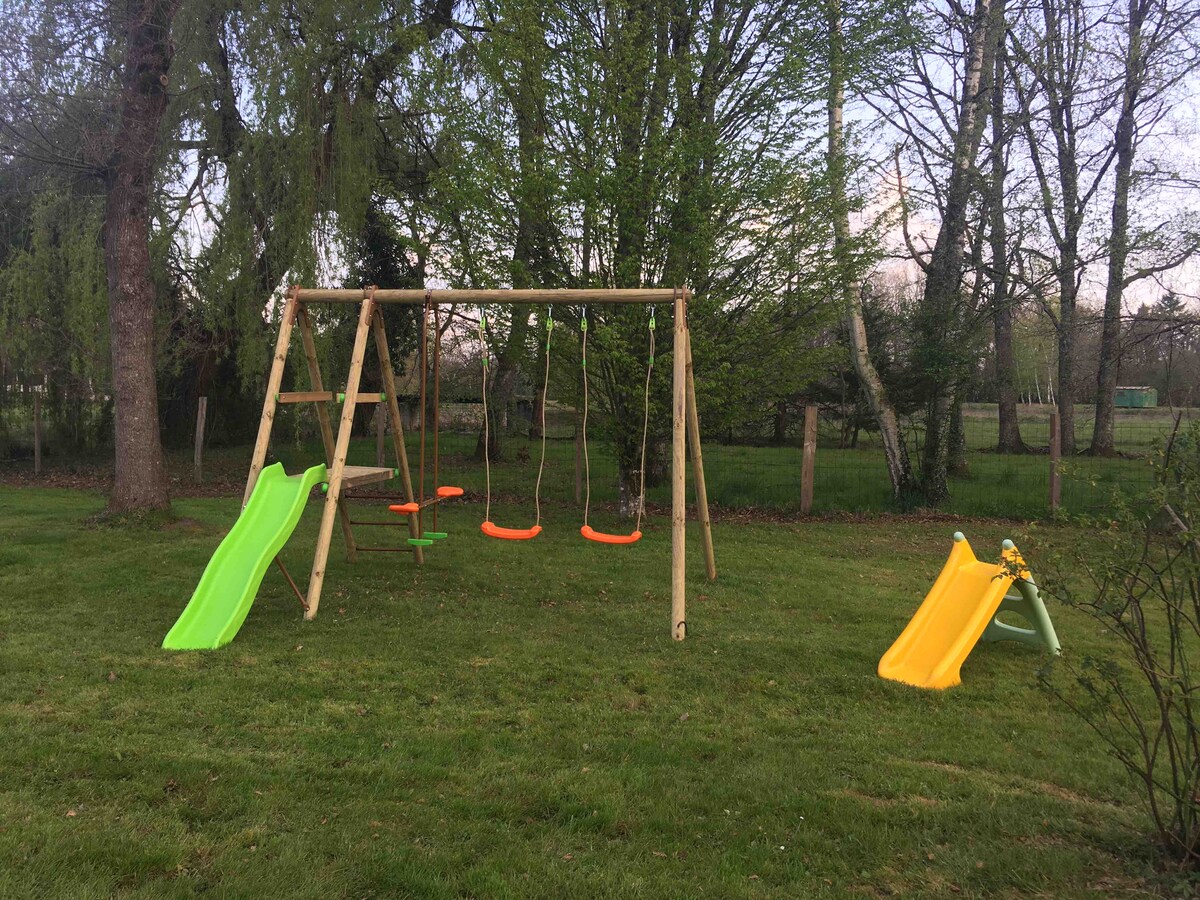 An outdoor play area features a wooden swing set with two swings in bright colors, surrounded by a grassy space. Two slides, one green and one orange, are positioned nearby, complemented by lush greenery and trees in the background.