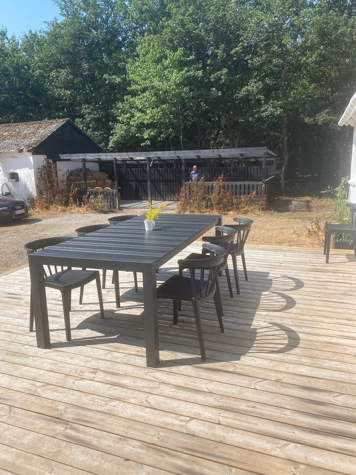 An outdoor terrace is shown featuring a large black dining table surrounded by six chairs. A small vase with yellow flowers is placed in the center of the table. In the background, greenery and an outbuilding are visible, providing a natural and serene setting.