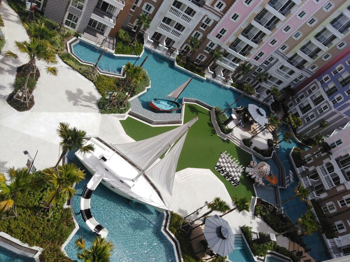 The image showcases a vibrant pool area at the condo complex, featuring a central sailing boat and green outdoor space. Surrounding palm trees and lounge chairs enhance the inviting ambiance, while the buildings' colorful facades create a cheerful backdrop.