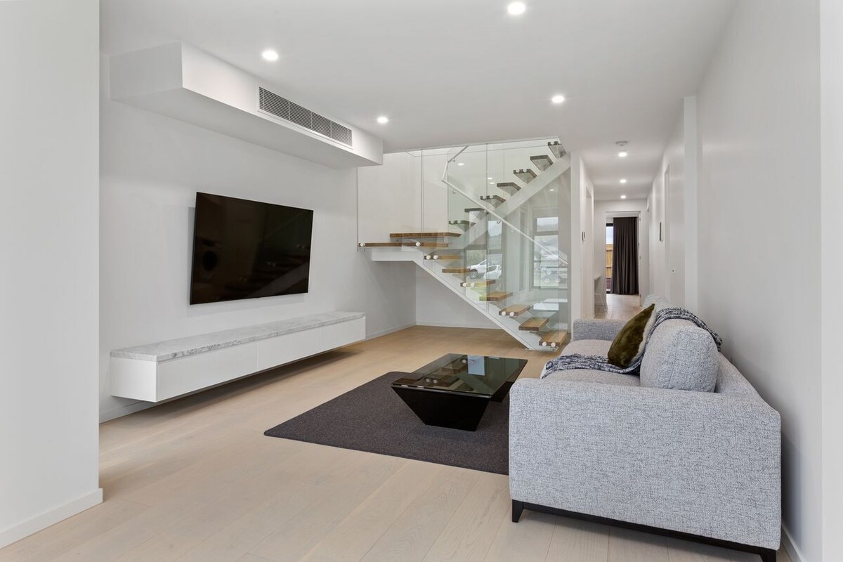 A spacious living area features a large flat-screen TV mounted on the wall above a sleek, white media console. A modern gray couch is accompanied by a glass coffee table, placed atop a soft area rug. Natural light fills the space from the staircase and adjacent areas.