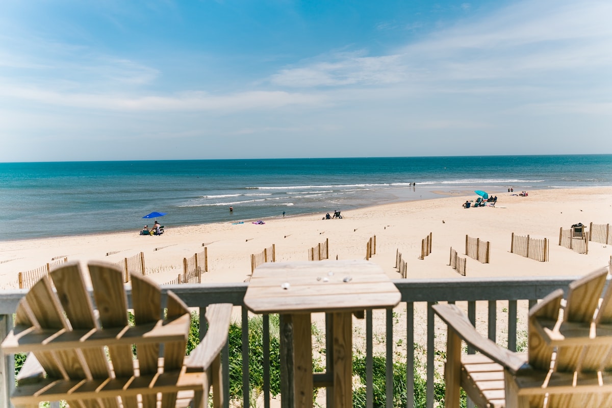 A serene beach view is visible from the porch, with soft sand and gentle waves in the distance. Beachgoers are seen engaging in various activities, and colorful umbrellas dot the shoreline. The outdoor seating provides a perfect spot for enjoying the ocean breeze.