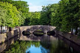 A stone bridge with three arches spans over a calm body of water. Lush green trees line the banks, their reflections visible on the water's surface, creating a serene scene set against a clear blue sky.