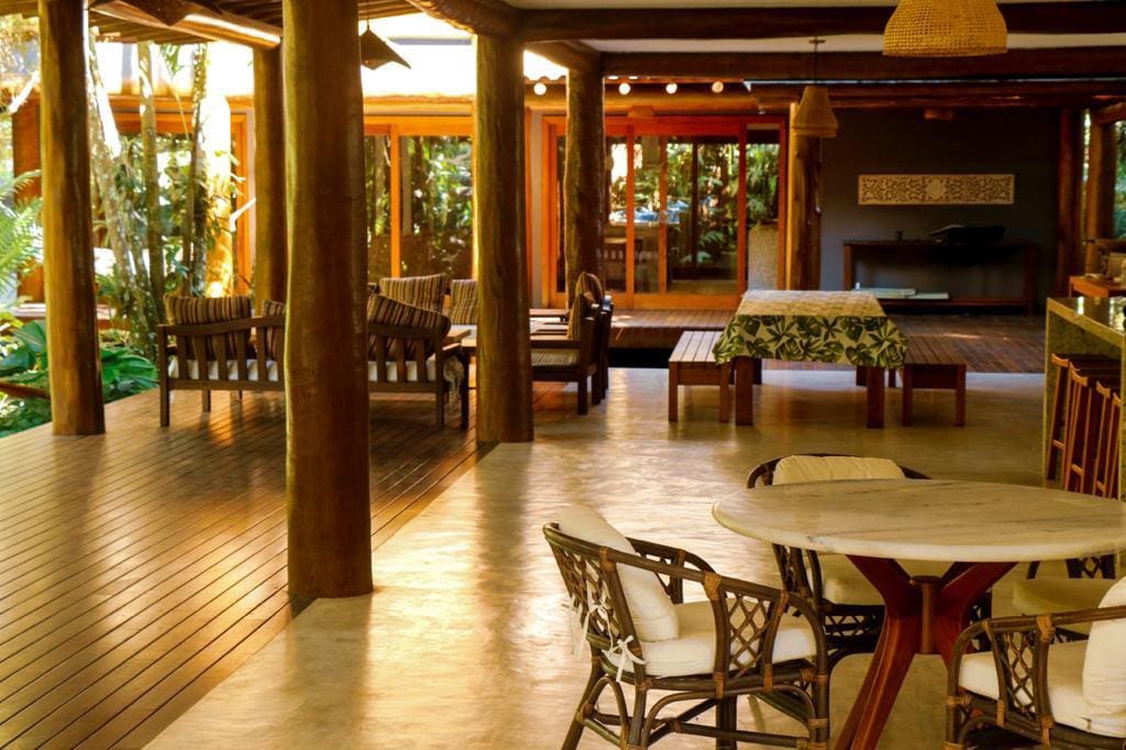 A spacious common area is highlighted by wooden beams and pillars. Comfortable seating arrangements are visible, including a large table with a patterned tablecloth. Natural light floods through expansive glass doors, revealing lush greenery outside.