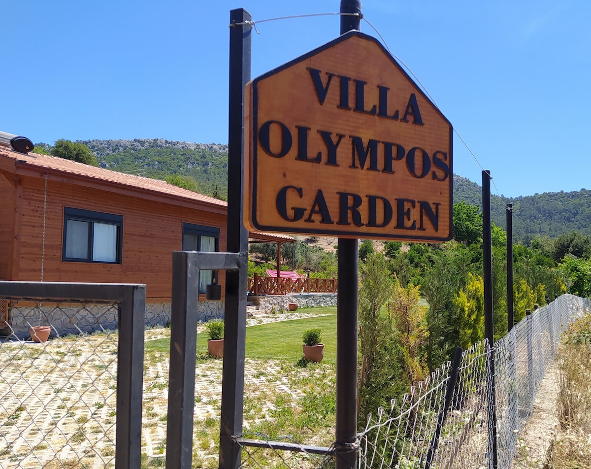 A wooden sign displays 'Villa Olympos Garden,' positioned at the entrance of a landscaped property. In the background, a modern villa with large windows is visible, surrounded by green grass and shrubbery. A wire fence defines the boundary, enhancing the peaceful setting.