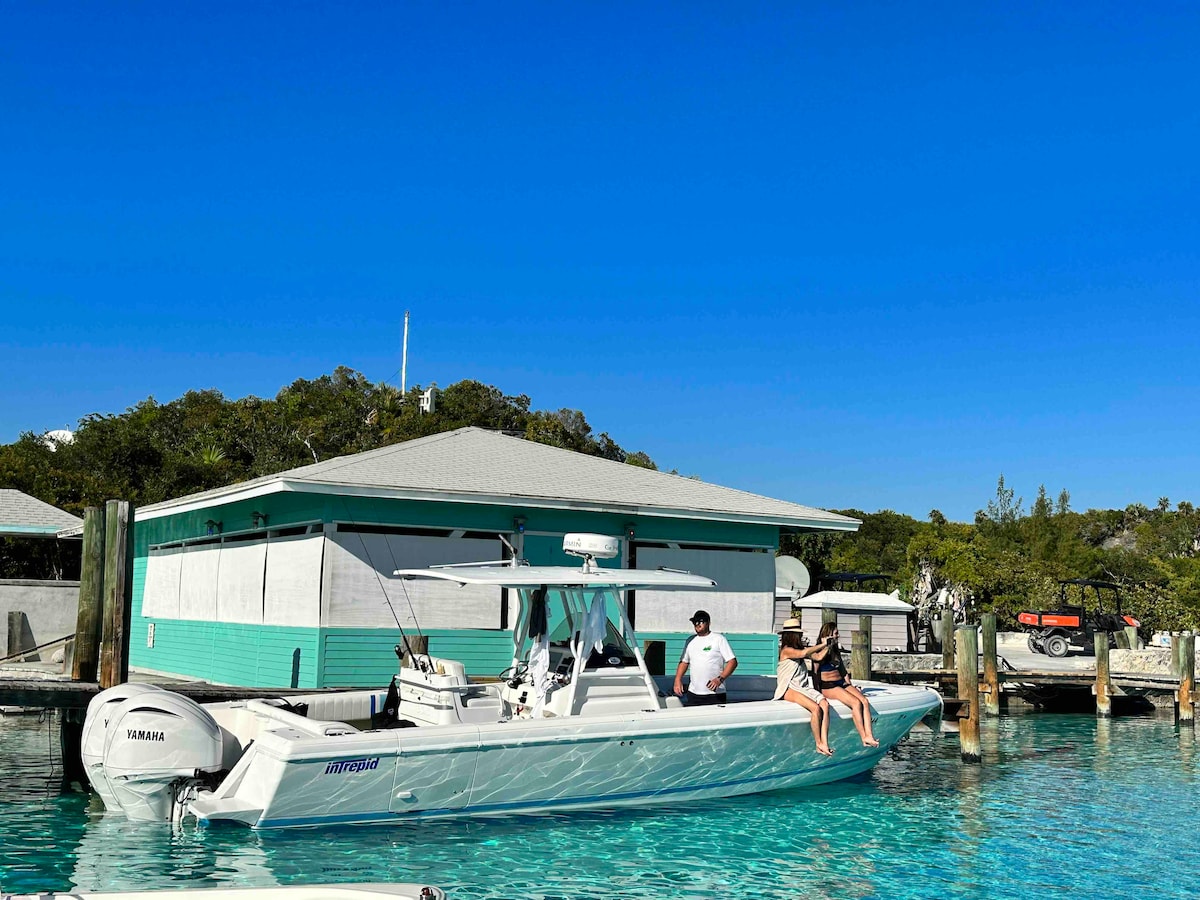 A motorboat is anchored in clear turquoise waters near a light green cottage. Two individuals are seated on the side of the boat, with the rear motor visible. The backdrop features lush greenery, and a blue sky is overhead.