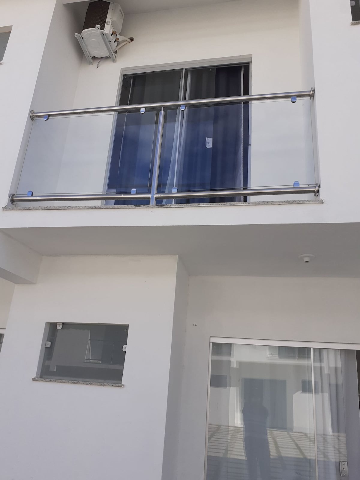 A modern balcony with a transparent glass railing is visible, providing unobstructed views. Large glass doors are seen leading to an interior space, allowing natural light to enter. The clean lines and white exterior create a fresh and open appearance.