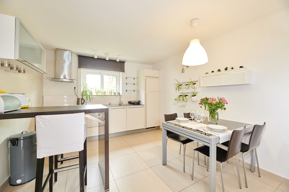 A modern kitchen and dining area is presented, featuring a spacious table set for six with floral centerpiece. Fully equipped cabinets and appliances are visible, alongside a sleek countertop. Natural light fills the room through the window, highlighting the neutral tones and functional design.