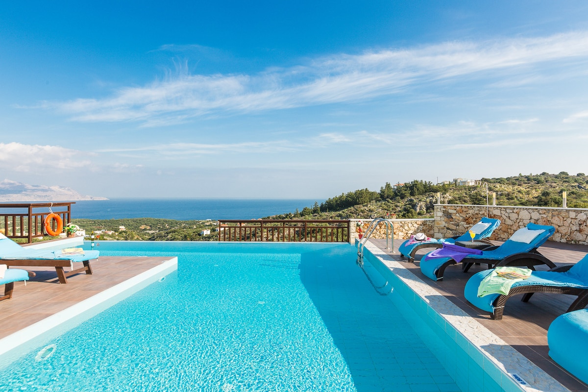 A private pool invites relaxation, surrounded by sun loungers. A scenic view of the sea and coastline is visible in the background. The pool area is complemented by a wooden railing, enhancing the open atmosphere.