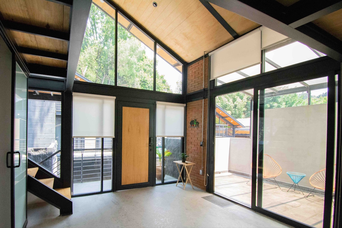 Natural light fills the spacious living area, characterized by tall windows and a wooden ceiling. Modern furniture includes a small table and two accent chairs, while a sliding door opens to a private outdoor patio. Greenery is visible outside, enhancing the serene atmosphere.