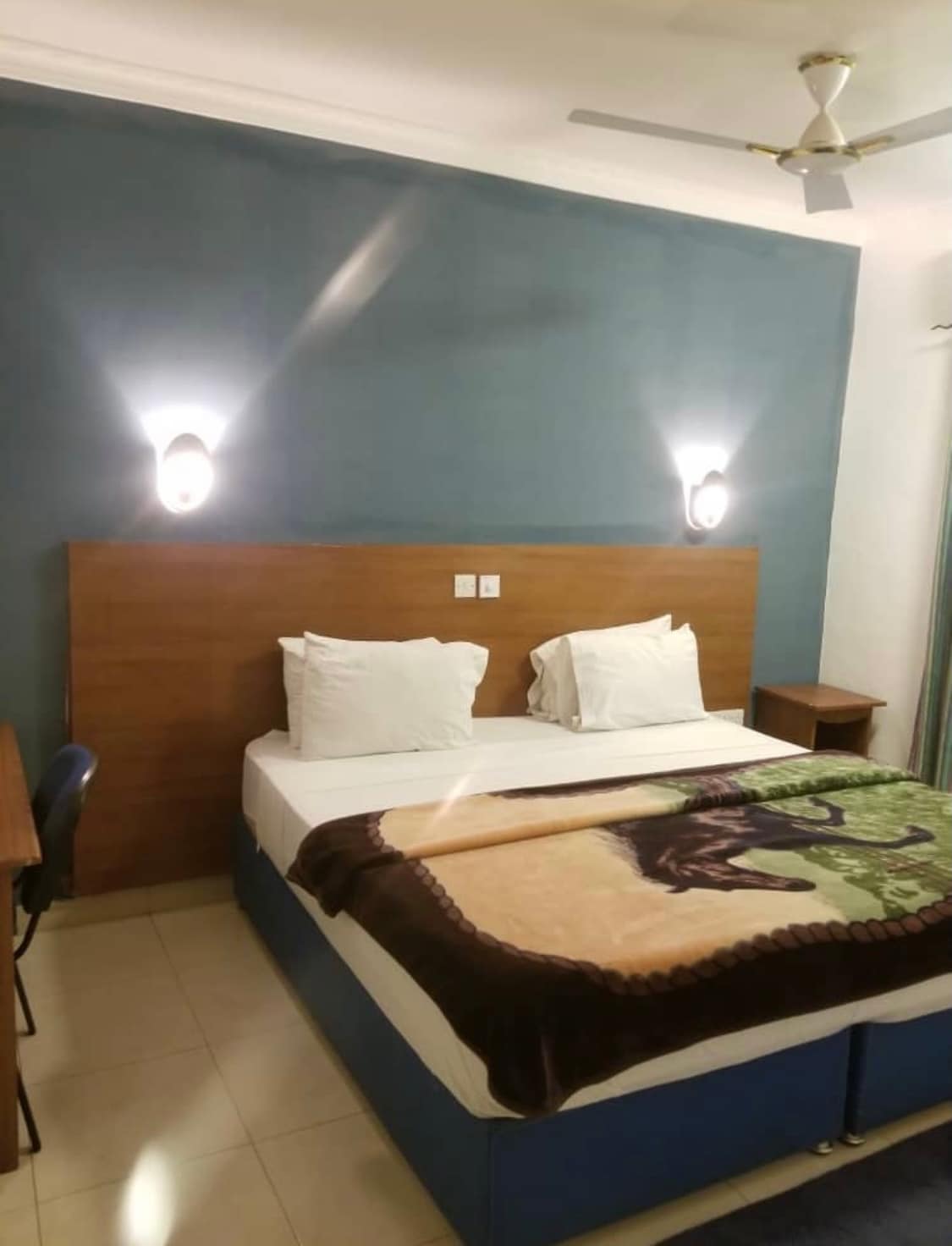 A comfortable room features a spacious bed with a colorful blanket draped across the foot. Soft lighting is provided by two wall-mounted fixtures. A wooden desk and chair are present, alongside a side table, enhancing the functionality of the space.