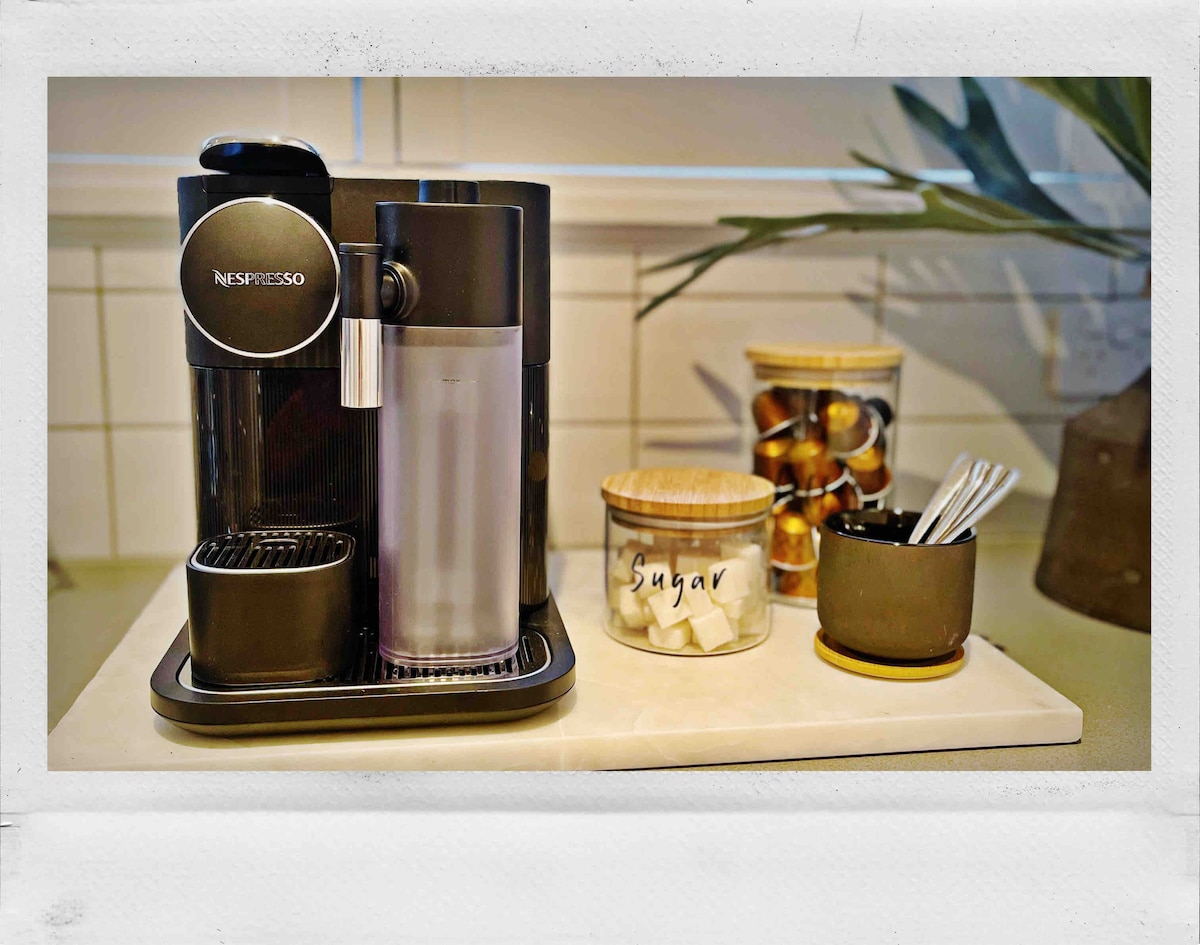A coffee station features a Nespresso machine beside a glass container holding coffee capsules. A small jar labeled 'Sugar' contains sugar cubes, and a ceramic dish holds stirrers, all arranged atop a neutral-colored countertop.