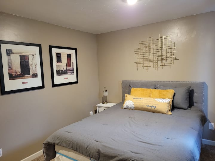 Nivel inferior quinto dormitorio (cama tamaño queen) recientemente decorado en un estilo unisex con un tema de bicicleta. 1 mesa auxiliar y una mesa más larga para tus artículos personales.  2 lámparas nuevas y nuevas mesas auxiliares  proporcionadas. 1 cómoda. Ropa de cama nueva y cómoda. Colores: gris y mostaza.